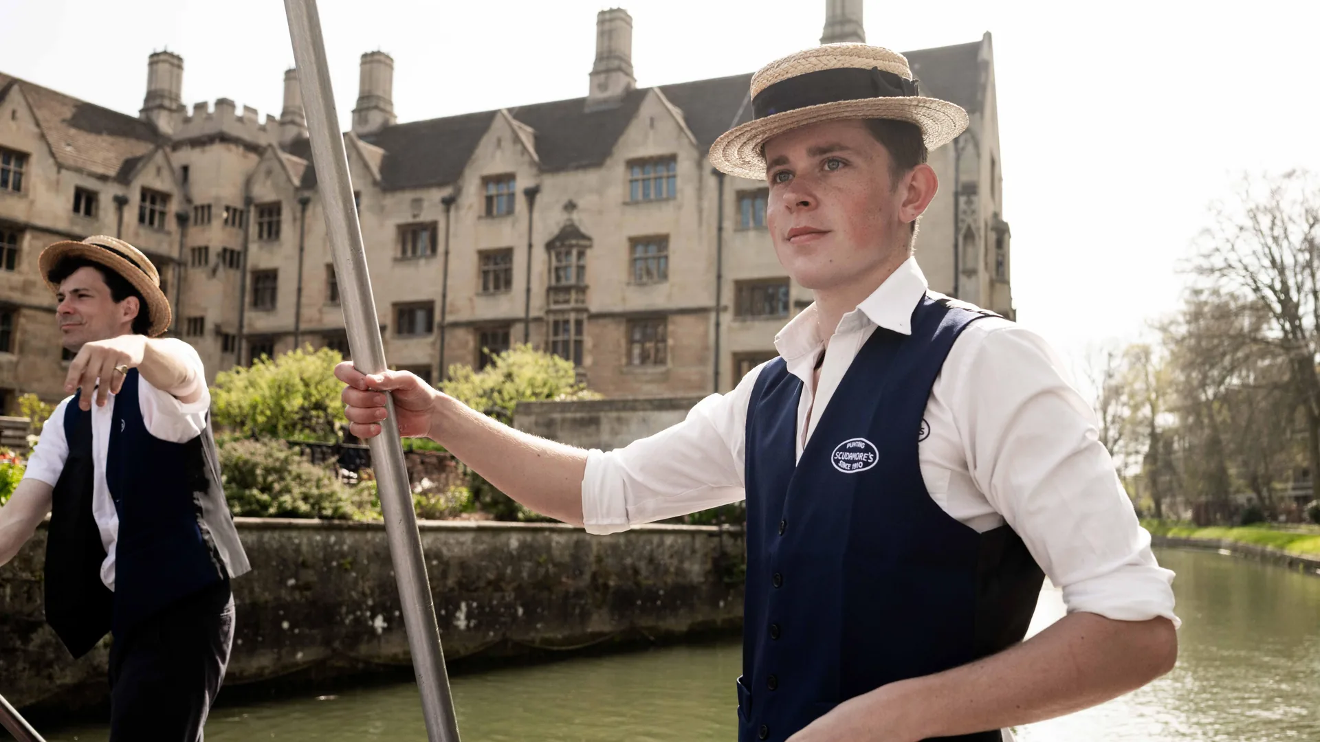 Two punt chauffeurs with poles beside a college on the River Cam, from the walk and punt tour listing